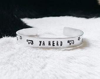 Op de afbeelding: Een zilveren armband met de woorden "YA HERD" gegraveerd. De armband is versierd met kleine koesilhouetten.