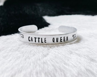 Może przedstawiać: Srebrna bransoletka z wygrawerowanym napisem "Cattle Queen". Po obu stronach tekstu znajdują się dwie małe sylwetki krów.