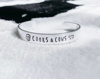 Może przedstawiać: Srebrna bransoletka metalowa z wygrawerowanym napisem "Coors & Cows". Na bransoletce wygrawerowano również kufel piwa i sylwetkę krowy.