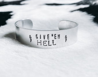 Op de afbeelding: Zilveren metalen armband met de tekst "GIVE 'EM HELL" en bliksemschicht symbolen aan beide kanten.