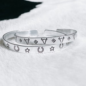 Puede incluir: Dos pulseras de plata con diseños de temática western. La pulsera superior presenta un patrón repetido de un cráneo de toro, un diamante y una estrella. La pulsera inferior presenta un patrón repetido de una herradura y una estrella.