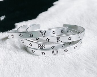 Op de afbeelding: Vier zilveren armbanden met verschillende ontwerpen in westernstijl. Elke armband heeft een rij sterren en een ander ontwerp, waaronder een cowboylaars, een stierenkop, een hoefijzer en een indiaans symbool.