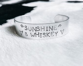 Op de afbeelding: Een zilverkleurige manchetarmband met de woorden "SUNSHINE & WHISKEY" gestempeld op de voorkant. De armband heeft een eenvoudig, open ontwerp en is versierd met kleine symbolen. De armband wordt gepresenteerd op een witte, getextureerde ondergrond.