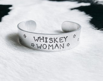 Op de afbeelding: Een zilveren metalen armband met de woorden "Whiskey Woman" erop gegraveerd. De armband is versierd met kleine sterretjes.