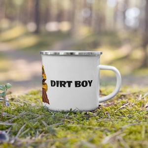 Könnte beinhalten: Weißer Emaillebecher mit silbernem Rand und Henkel. Der Becher trägt den Text "DIRT BOY" in fetten schwarzen Buchstaben. Auf der linken Seite des Bechers befindet sich eine farbenfrohe Grafik einer Person. Der Becher steht auf einem Bett aus grünem Moos.