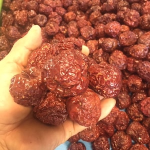May include: A handful of dried, reddish-brown jujube fruits, with a wrinkled texture, are held in a hand. The background is filled with more of the same dried fruit. The image highlights the color and texture of the jujubes.