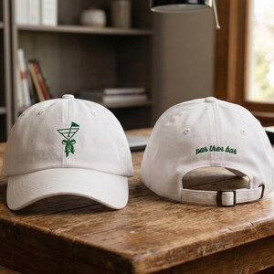 May include: Two white baseball caps with green embroidered designs. One cap features a cocktail glass with a golf flag, the other has "par then bar" embroidered on the back. Both caps are on a wooden table.