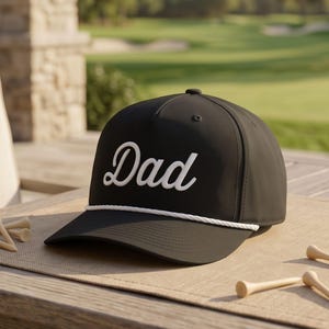 Puede incluir: Una gorra de b&eacute;isbol negra con la palabra Dad bordada en letra cursiva blanca en el panel frontal. La gorra incluye un detalle de cuerda blanca en la base de la corona, justo encima de la visera, y tiene un dise&ntilde;o estructurado de cinco paneles.
