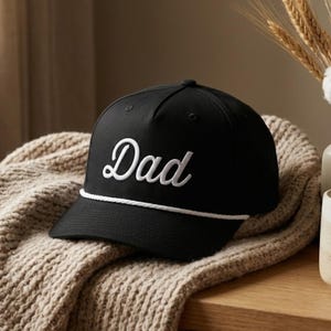 Puede incluir: Una gorra de b&eacute;isbol negra con la palabra Dad bordada en letra cursiva blanca en el panel frontal. La gorra incluye un detalle de cuerda blanca que recorre horizontalmente la base de la corona, justo encima de la visera curva y estructurada.