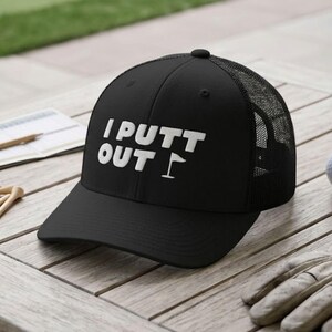 May include: A black trucker cap featuring the white text I PUTT OUT with a small golf flag icon. The cap has a solid front panel and a mesh back. It is displayed alongside three white golf balls, a beige golf glove, and wooden golf tees.
