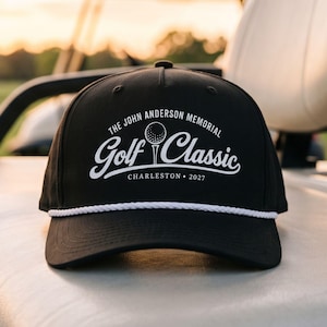 May include: A black baseball cap featuring a white rope detail across the base of the crown. The front displays white text reading THE JOHN ANDERSON MEMORIAL GOLF CLASSIC CHARLESTON 2027 with a graphic of a golf ball on a tee in the center.