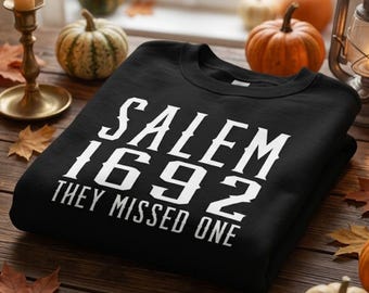May include: A black crewneck sweatshirt featuring white text that reads SALEM 1692 THEY MISSED ONE. The garment is displayed on a wooden surface surrounded by various small pumpkins and autumn leaves, with a lit candle and a lantern nearby.