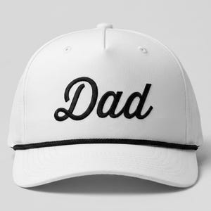 Puede incluir: Una gorra de b&eacute;isbol blanca con la palabra Dad bordada en una fuente cursiva negra en el panel frontal. La gorra incluye un detalle de cuerda negra que recorre horizontalmente la base de la corona, justo encima de la visera curva y estructurada.