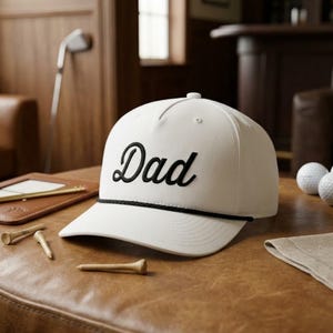 Puede incluir: Una gorra de b&eacute;isbol blanca con la palabra Dad bordada en letras cursivas negras en el panel frontal. La gorra incluye un detalle de cuerda negra que recorre horizontalmente la base de la corona, justo encima de la visera curva.