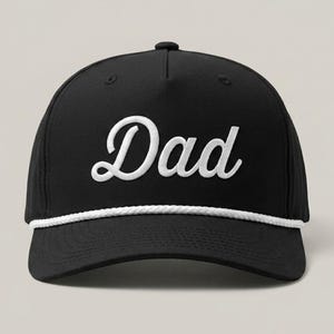 Puede incluir: Una gorra de b&eacute;isbol negra con la palabra Dad bordada en fuente cursiva blanca en el panel frontal. La gorra incluye un detalle de cuerda trenzada blanca que recorre horizontalmente la base de la corona, justo encima de la visera curva.