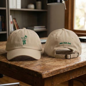 May include: Two tan baseball caps. One cap features a green embroidered cocktail glass design on the front. The other cap has the words "par then bar" embroidered in green on the back. Both caps have adjustable straps.