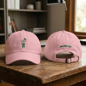 May include: Two pink baseball caps on a wooden surface. The front cap has a green embroidered design of a martini glass with a golf flag. The back cap has the words "par then bar" embroidered in green.