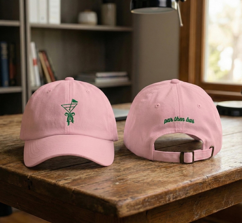 May include: Two pink baseball caps on a wooden surface. The front cap has a green embroidered design of a martini glass with a golf flag. The back cap has the words "par then bar" embroidered in green.