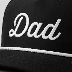 Puede incluir: Una gorra de b&eacute;isbol negra con la palabra Dad bordada en letra cursiva blanca en el panel frontal. La gorra incluye un detalle de cord&oacute;n trenzado blanco colocado horizontalmente en la base de la corona, justo encima de la visera.