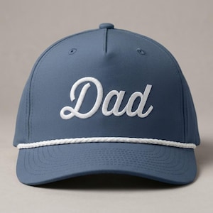 Puede incluir: Una gorra de b&eacute;isbol color azul pizarra con la palabra Dad bordada en letra cursiva blanca en el panel frontal. La gorra incluye un detalle de cuerda trenzada blanca que recorre horizontalmente la base de la corona, sobre la visera curva.