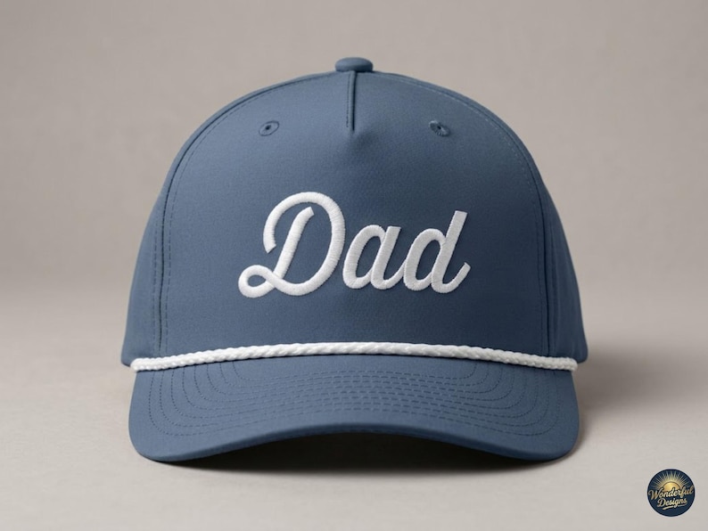 Puede incluir: Una gorra de b&eacute;isbol color azul pizarra con la palabra Dad bordada en letra cursiva blanca en el panel frontal. La gorra incluye un detalle de cuerda trenzada blanca que recorre horizontalmente la base de la corona, sobre la visera curva.