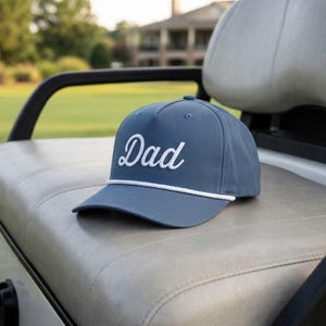 Puede incluir: Una gorra de b&eacute;isbol color azul pizarra con la palabra Dad bordada en fuente cursiva blanca en el panel frontal. La gorra incluye un detalle de cuerda trenzada blanca que recorre horizontalmente la base de la corona, sobre la visera curva.
