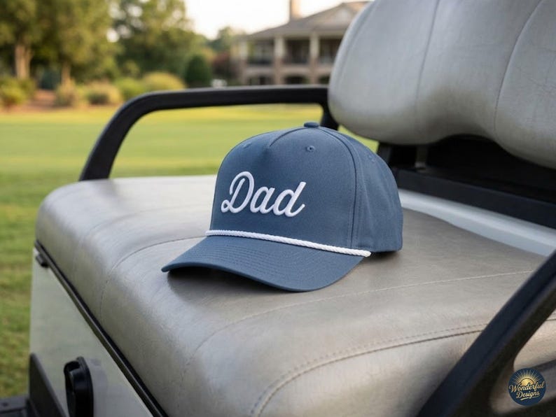 Puede incluir: Una gorra de b&eacute;isbol color azul pizarra con la palabra Dad bordada en fuente cursiva blanca en el panel frontal. La gorra incluye un detalle de cuerda trenzada blanca que recorre horizontalmente la base de la corona, sobre la visera curva.