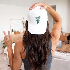 May include: A white baseball cap with a green cocktail glass design. The cap is made of a soft material and has a curved brim. The design is embroidered on the front of the cap. The cap is being held up by a person.