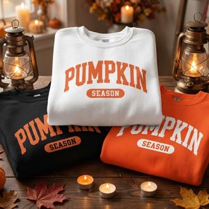 May include: Three crewneck sweatshirts in white, black, and orange. Each features the text PUMPKIN SEASON in a bold, arched font. The white sweatshirt has orange text, the black has orange text, and the orange sweatshirt has white text.
