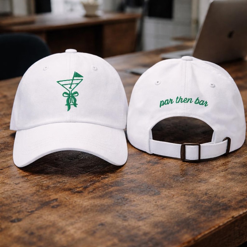 May include: Two white baseball caps. The front cap has a green embroidered design of a golf flag in a martini glass. The back cap has the words "par then bar" embroidered in green. Both caps have an adjustable strap.
