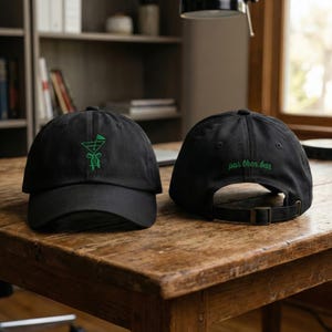 May include: Two black baseball caps on a wooden table. One cap features a green embroidered cocktail design, the other has the text "par then bar" in green. Both caps have adjustable straps.