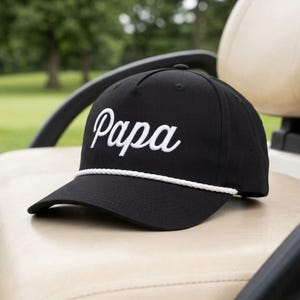 May include: A black baseball cap featuring the word Papa embroidered in white script font on the front panel. The cap includes a white braided rope detail across the base of the crown, just above the curved brim, and has a structured five-panel design.