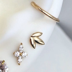 May include: Gold ear piercings with clear gemstones and a gold leaf design. The piercings are on a white mannequin head.