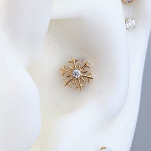 Peut inclure: Boucles d'oreilles en forme de flocon de neige dorées avec des pierres précieuses transparentes. Les boucles d'oreilles sont présentées sur une tête de mannequin blanche avec plusieurs piercings.