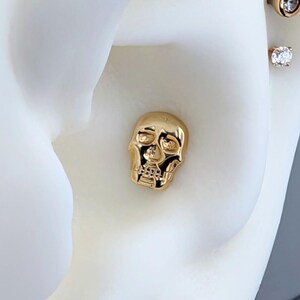 May include: A collection of gold-toned earrings displayed on a white ear model. The earrings include a skull stud, a round stud, a bezel-set diamond, a small opal stud, a star-shaped diamond stud, and a large round diamond stud.