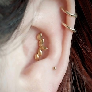 May include: Close-up of an ear adorned with gold jewelry. A curved, leaf-shaped earring is set in the helix, with two gold hoops and a small stud. The ear is set against a backdrop of auburn hair.