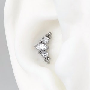 Può includere: Un orecchino a cuffia in argento con tre pietre preziose trasparenti a forma di marquise e piccole perle d'argento. Il cuffia è indossato sul cartilagine superiore dell'orecchio.