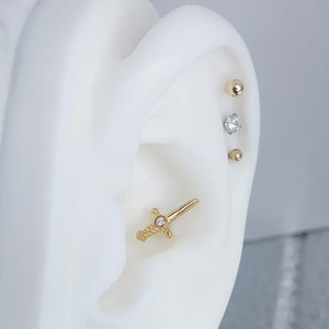 Pode incluir: Piercing de cartilage da orelha em ouro com um pequeno diamante, uma bola dourada e um pequeno diamante. O piercing tem a forma de uma espada.