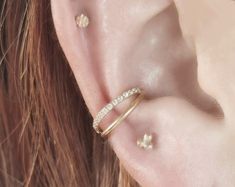 May include: A close-up of a person's ear with multiple gold earrings. The earrings include a small gold stud, a larger gold stud with a small diamond, a gold hoop with a row of small diamonds, and a gold hoop with a small star-shaped charm.