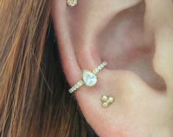 Peut inclure: Gros plan sur l'oreille d'une personne avec plusieurs piercings. L'oreille est percée avec une boucle d'oreille en or avec un diamant en forme de larme, un petit clou en or et un clou plus grand avec une pierre transparente.