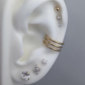 May include: An ear with multiple piercings, showcasing various stud earrings and a triple-band ear cuff. The studs include a gold ball, clear crystals, and a flower-shaped design. A small, square opal stud is also visible.