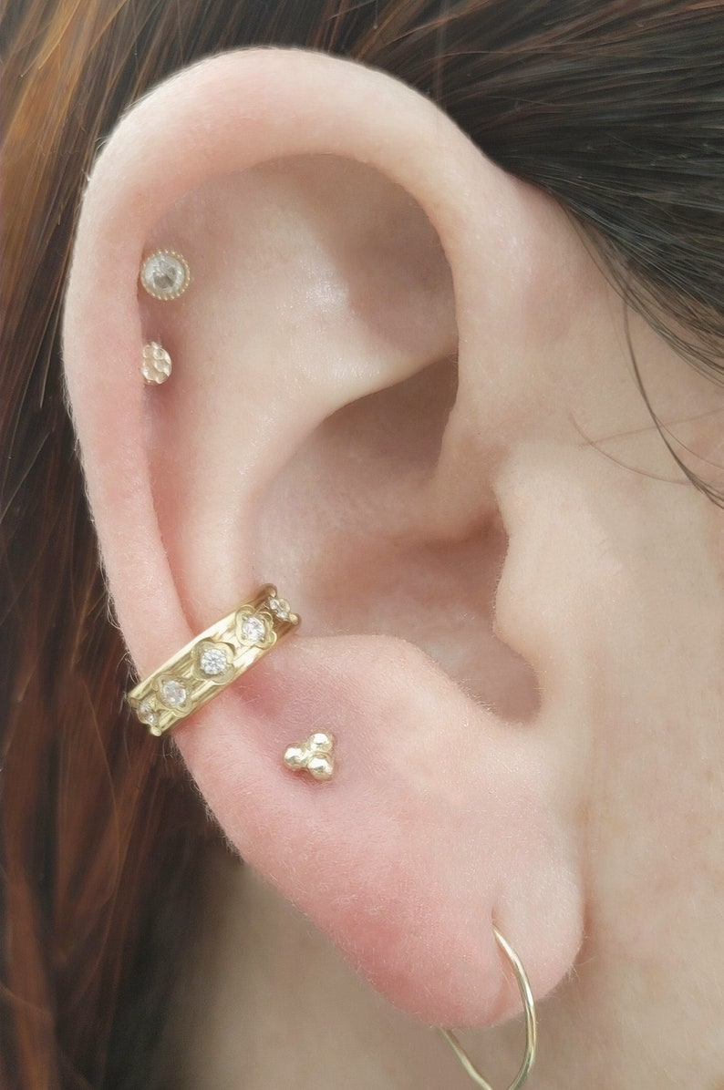 K&ouml;nnte beinhalten: Ein Ohr mit mehreren Piercings. Das Ohr ist mit einem goldenen Creolenohrring mit kleinen Diamanten, einem goldenen Ohrstecker mit einem kleinen Diamanten, einem goldenen Ohrstecker mit einem strukturierten Design und einem goldenen Ohrstecker mit drei kleinen Kugeln gepierct.