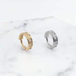 K&ouml;nnte beinhalten: Zwei Creolen mit einem dekorativen Design mit kleinen, klaren Steinen. Die Creole links ist golden und die Creole rechts ist silber.