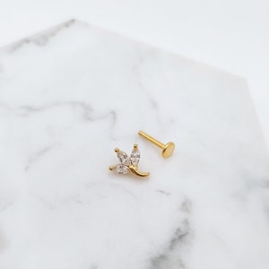 Puede incluir: Joyer&iacute;a de cuerpo dorada con un peque&ntilde;o dise&ntilde;o de flor de cristal transparente. La joyer&iacute;a es un pendiente con la parte posterior plana.