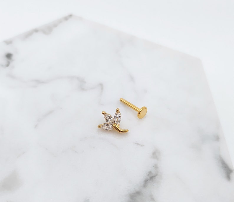 Puede incluir: Joyer&iacute;a de cuerpo dorada con un peque&ntilde;o dise&ntilde;o de flor de cristal transparente. La joyer&iacute;a es un pendiente con la parte posterior plana.