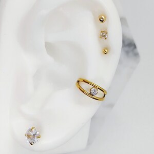 May include: Gold ear cuff with a single clear rhinestone in the center. The cuff is made of two thin bands of metal that are connected at the top and bottom. The cuff is worn on the upper part of the ear. There are also three gold studs with clear rhinestones in the earlobe.