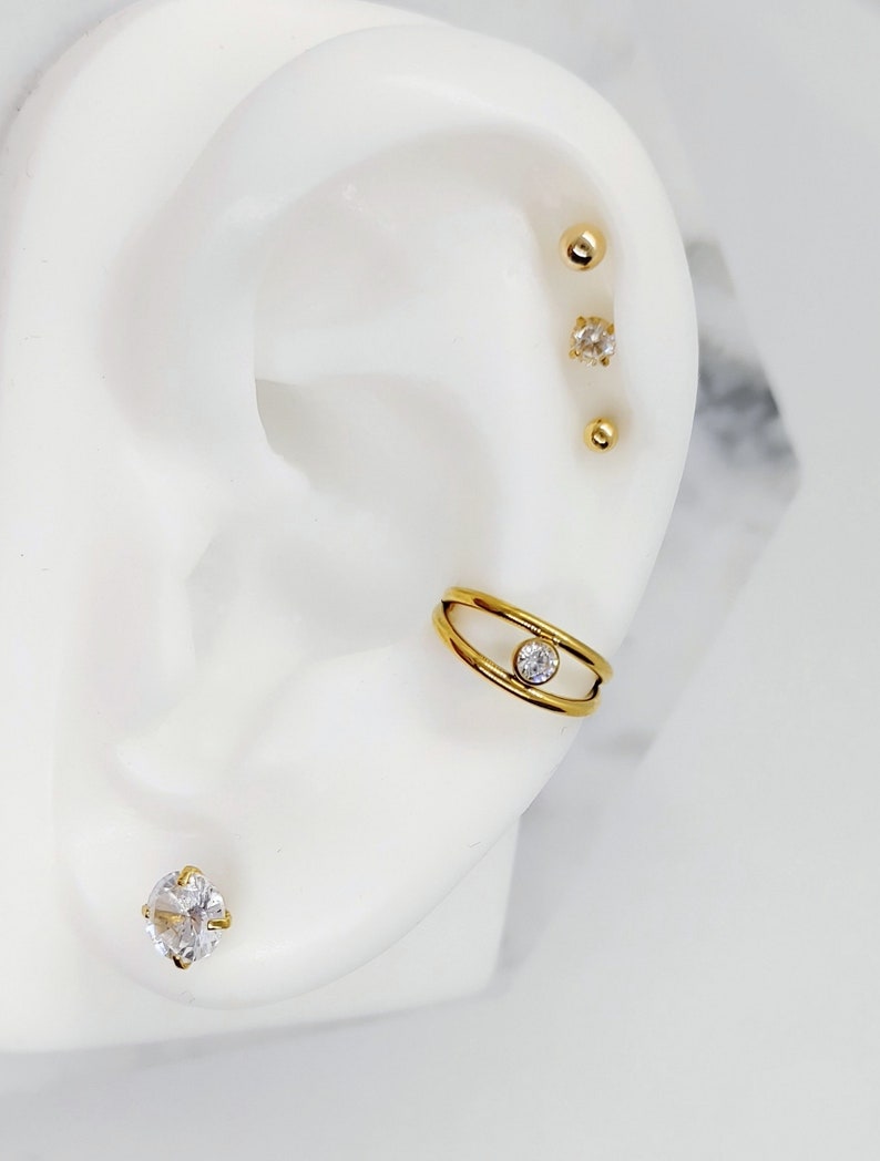 May include: Gold ear cuff with a single clear rhinestone in the center. The cuff is made of two thin bands of metal that are connected at the top and bottom. The cuff is worn on the upper part of the ear. There are also three gold studs with clear rhinestones in the earlobe.