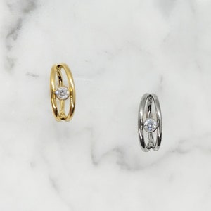 May include: Two nose rings with a single clear stone in the center. The ring on the left is gold and the ring on the right is silver.