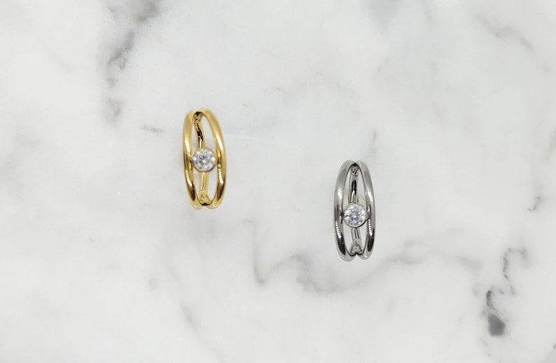 May include: Two nose rings with a single clear stone in the center. The ring on the left is gold and the ring on the right is silver.