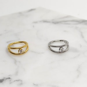 May include: Two simple rings with a single clear stone set in the center. The ring on the left is gold and the ring on the right is silver.